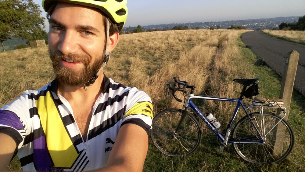 A man in a yellow helmet takes a selfie with his bike and a field of grass in the background.