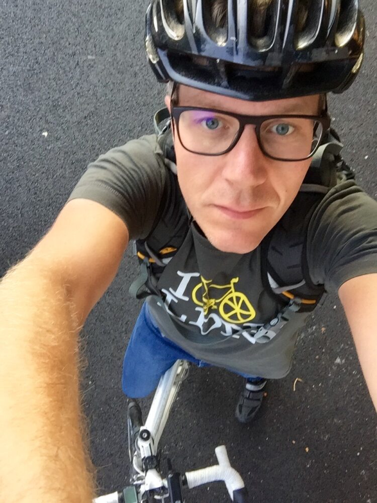 A man on a bike wearing glasses and a bike helmet takes a selfie from above.