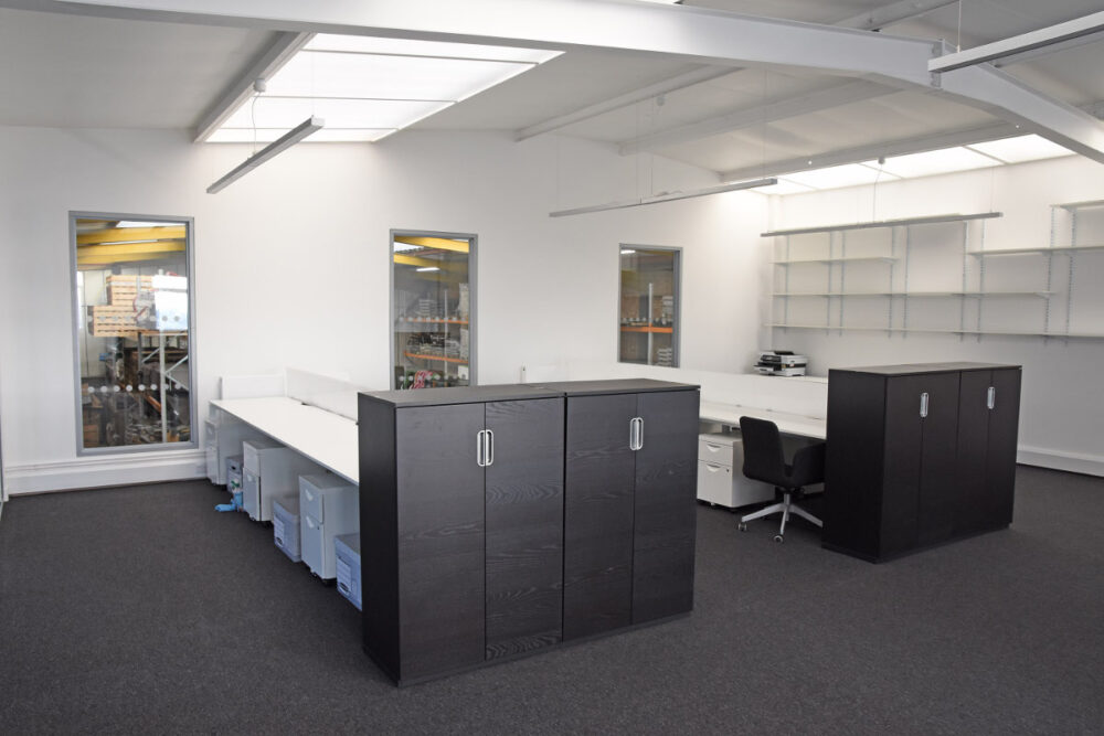 An empty office with white desks, grey carpet and black storage cupboards.