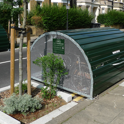 A dome-roofed Bikehangar storage container with a perforated metal side and a green metal roof, next to a tree pit beside a footpath.