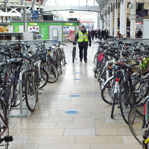 A person in a high-vis vest wheels a bike towards a group of bikes parked against metal bike racks at Paddington Station.