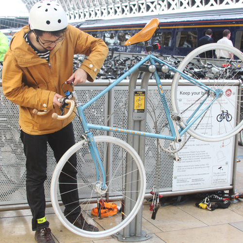 A man wearing a helmet and a yellow jacket works on the handlebars of his bike, which is suspended by a public bike repair stand.