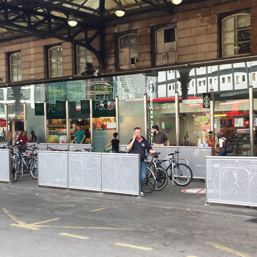 People walk through and past a busy bike parking area marked by metal railings outside a train station.