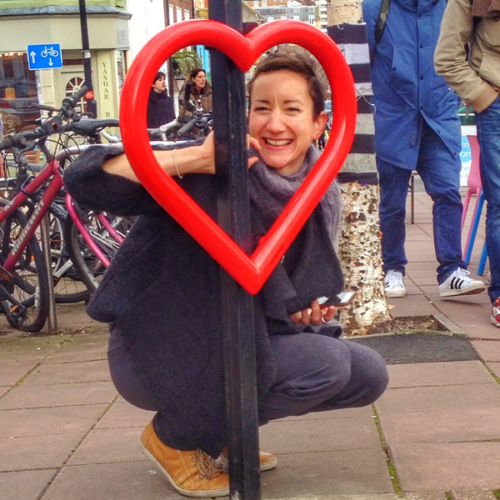 A woman crouches down on the street and smiles through a red, heart-shaped Lovehoop bike stand affixed to a light post.