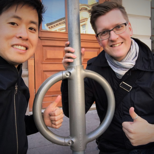 Two people smile and give a thumbs up to the camera either side of a metal Cyclehoop bike stand.