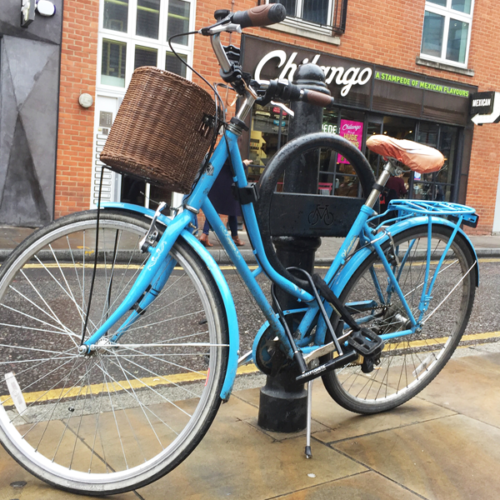 A blue bicycle parked against a black Cyclehoop bike rack affixed to a bollard on an urban street.