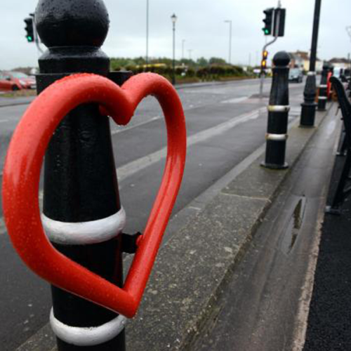 A red heart-shaped Lovehoop bike stand, attached to a bollard on the side of a road.