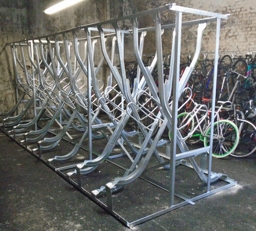 An empty metal rack for vertical bike storage, with a full rack behind it, both inside a concreted room.