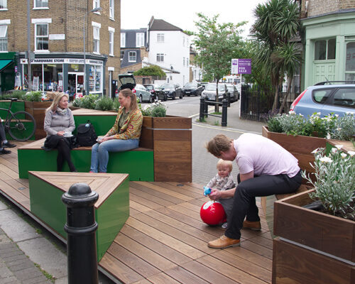 People sit and chat on benches in a street car park that has been converted into wooden seating with greenery.