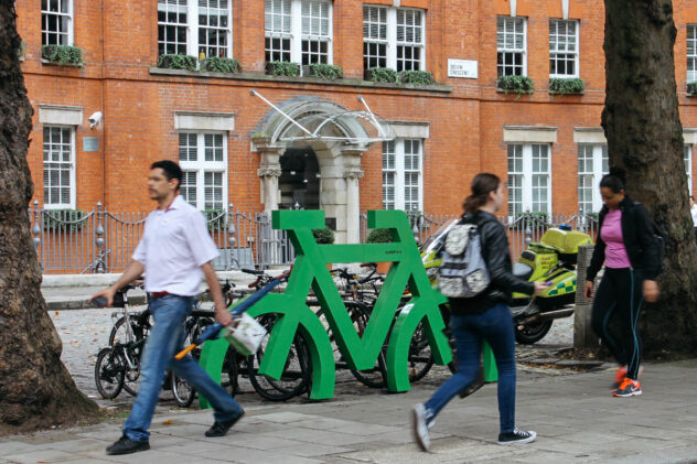 People walk past a green Bike Port bike storage rack shaped like a bicycle, with bikes parked against it.