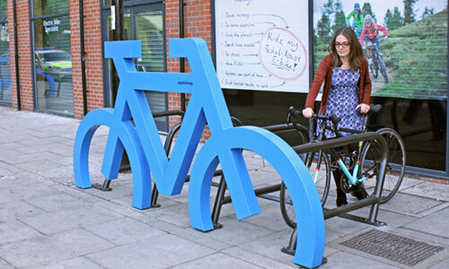 A woman parks her bike in a large blue Cyclehoop bike port in the shape of a bicycle.