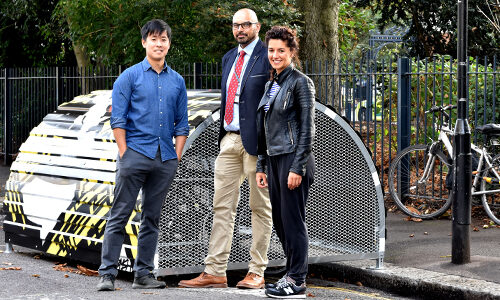 Three people stand in front of a some-roofed Bikehangar bike storage container on the side of a road.