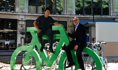 Two men sit on a green Bike Port bike storage rack shaped like a bicycle, against which bicycles are parked.