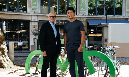 Two men stand in front of a green Bike Port bike storage rack in the shape of a bicycle.