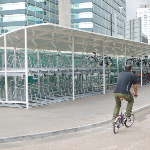 A man rides a foldable bike past a double-layered bike storage rack with skyscrapers in the background.