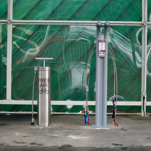 A metal bike pump and a taller metal public bike repair stand with various tools attached to it by wire rope, both bolted to a concrete floor.