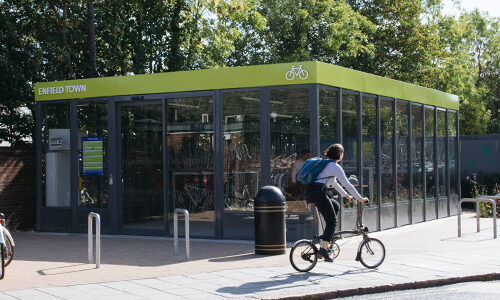 A person rides a foldable bike past the Bike Hub at Enfield Town, a glass-walled bike storage facility with a green roof.
