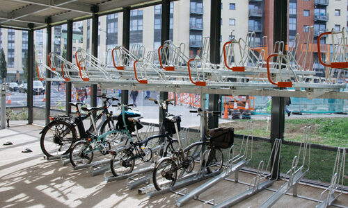 Bikes parked on the lower layer of a two-tier metal bike rack in a glass-walled structure overlooking a building site.