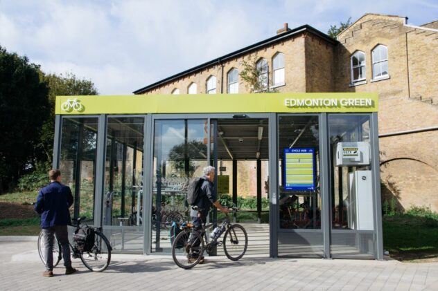 Two people stand with bikes outside the Bike Hub at Edmonton Green, a glass-walled bike storage facility with a green roof.