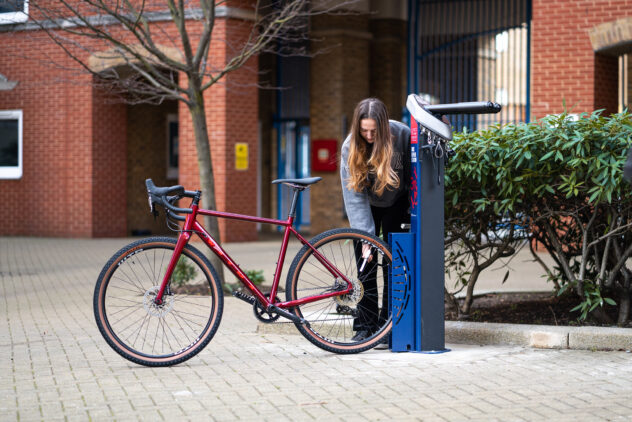 A woman pumps up the rear tire of her red bike using a blue Public Bike Stand in the middle of a cobblestone square.