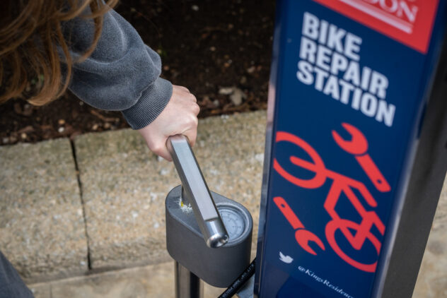 A person holds the handle of a Cyclehoop Deluce Repair Stand pump next to a sign that says Bike Repair Station.