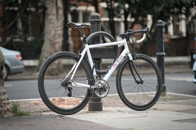 A white bike leaning against a street bollard, attached to a circular metal Cyclehoop.