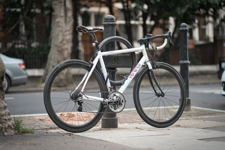 A white bike leaning against a street bollard, attached to a circular metal Cyclehoop.