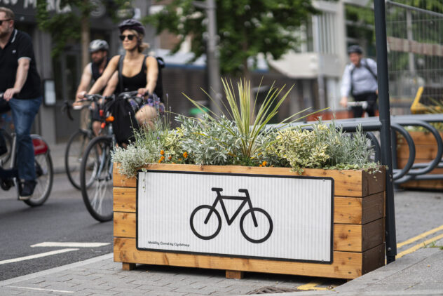 A wooden planter box with a large bicycle icon on its side, with cyclists riding past in the background.