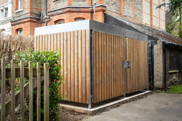 A lockable rectangular bike shed wood-paneled walls, in front of a stately old brick building.