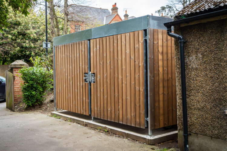 A rectangular panelled Wood Bike Shelter next to a house with a driveway.