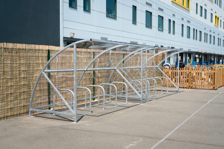 A row of metal bike storage racks under a clear canopy against a wooden fence.