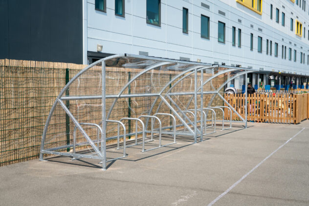 A row of metal bike storage racks under a clear canopy against a wooden fence.