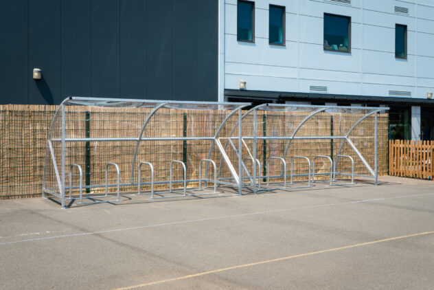 A row of metal bike stands underneath a clear canopy against a wooden fence.