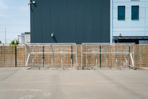 A metal bike rack in a concrete open air carpark against a wooden fence.