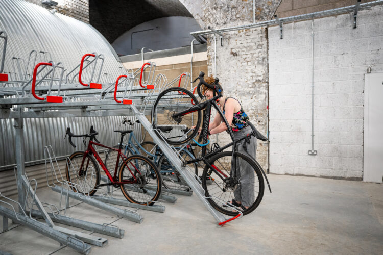 A woman wheels her bike up a ramp onto the top layer of a two-tier metal bike rack inside a concrete room.