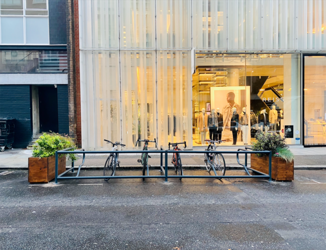 Bikes parked against metal rails between two wooden planters outside of a shopfront.