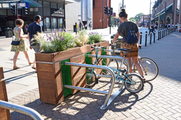 A woman parks her bike against a metal bike stand attached to a wooden planter box on the side of a paved street.