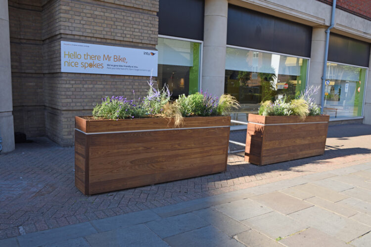 Two wooden planter boxes with plants inside on a street, in front of a wall that has a sign which reads Hello there Mr Bike, nice spokes.