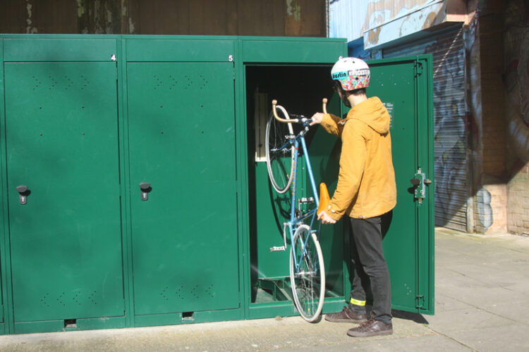 A person wearing a yellow jacket and a helmet places a bicycle into a green metal Vertical Bike Locker.