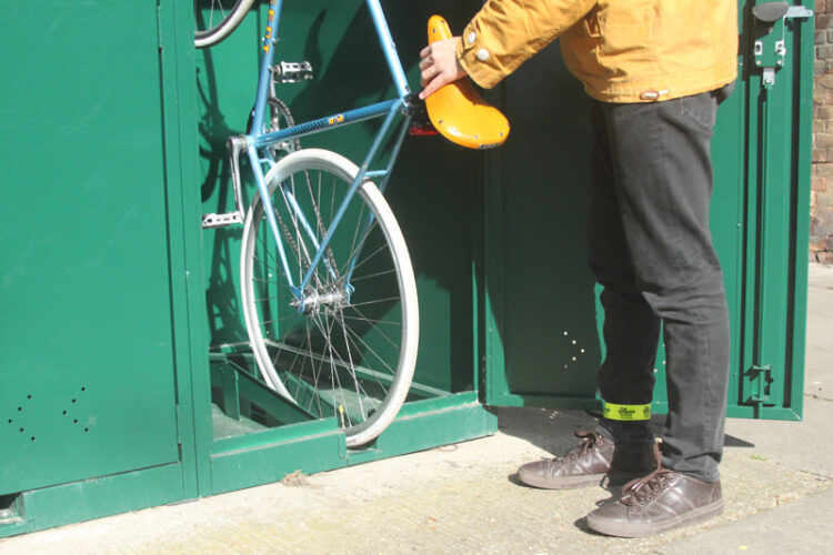 A person wearing a yellow jacket places a bicycle into a green metal Vertical Bike Locker.
