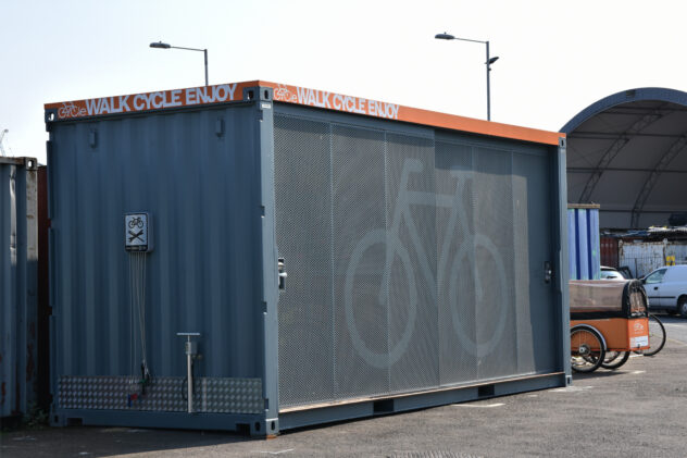 A Cyclehub container made of perforated gray metal with a bike symbol on the front and an orange painted awning with Walk Cycle Enjoy painted on in white.