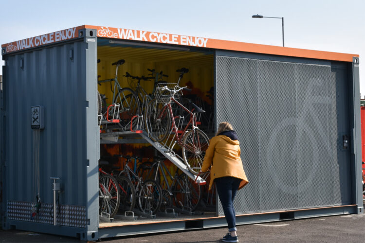 A person in a yellow coat wheels a bike up a ramp, into a large Cyclehoop storage container full of bikes.