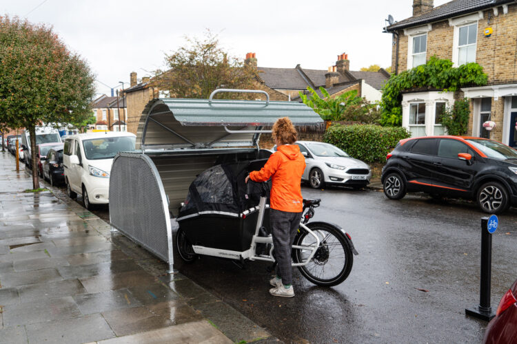 A person wearing an orange jacket parks their cargo bike inside a dome-roofed Bikehangar bike storage container on a residential street.