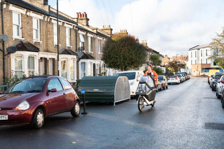 A woman rides a cargo bike with a pram attachment over the front wheel past a dome-roofed Bikehangar storage container on a residential street.