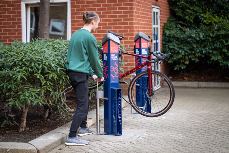 A man uses a bike tire pump at a public bike repair stand, from which his red bicycle is suspended.