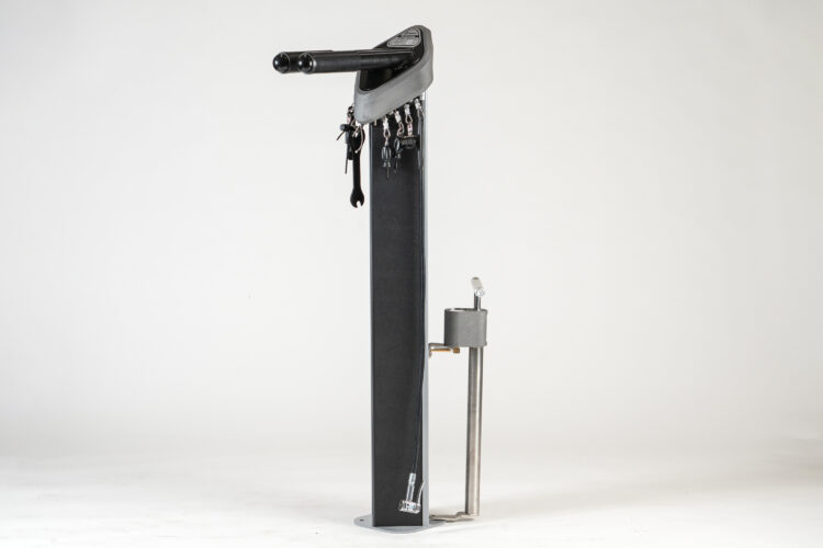A cylindrical metal bike repair station in black and silver metal, from which various tools hang.