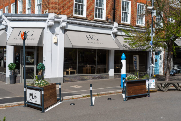 Two bike parking spaces marked with white lines on the road and a sign with an image of a cargo bike between two wooden planters in front of a cafe.