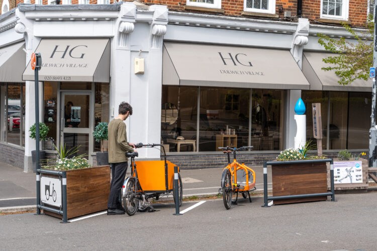 A man parks his orange cargo bike against a metal bike rack between two wooden planters outside of a cafe.