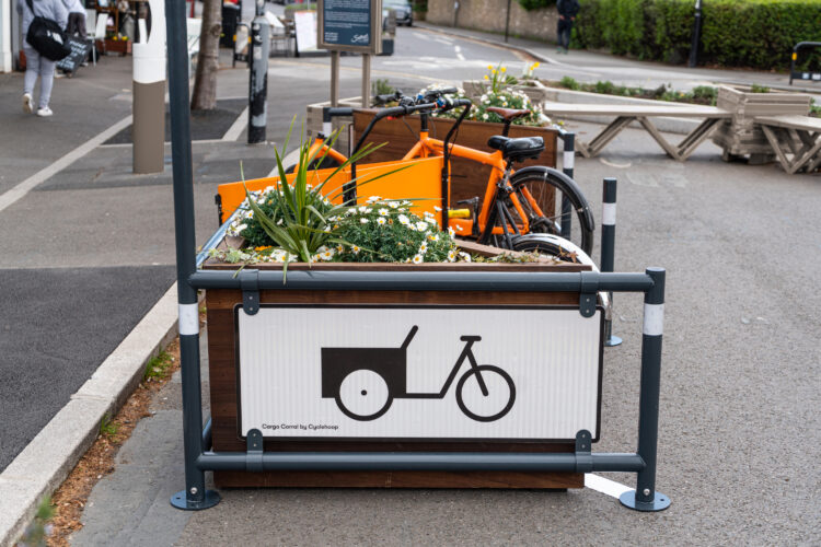 A sign with an image of a cargo bike painted on to it, on the edge of a bike parking space flanked by planter boxes.