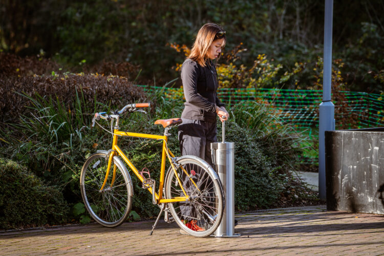 A woman pumps up her bike's rear tire at a metal Public Bike Repair Stand.